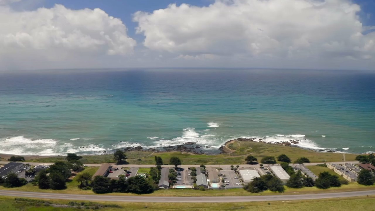 Aerial view of a hotel complex with a pool and parking lot, overlooking the ocean.