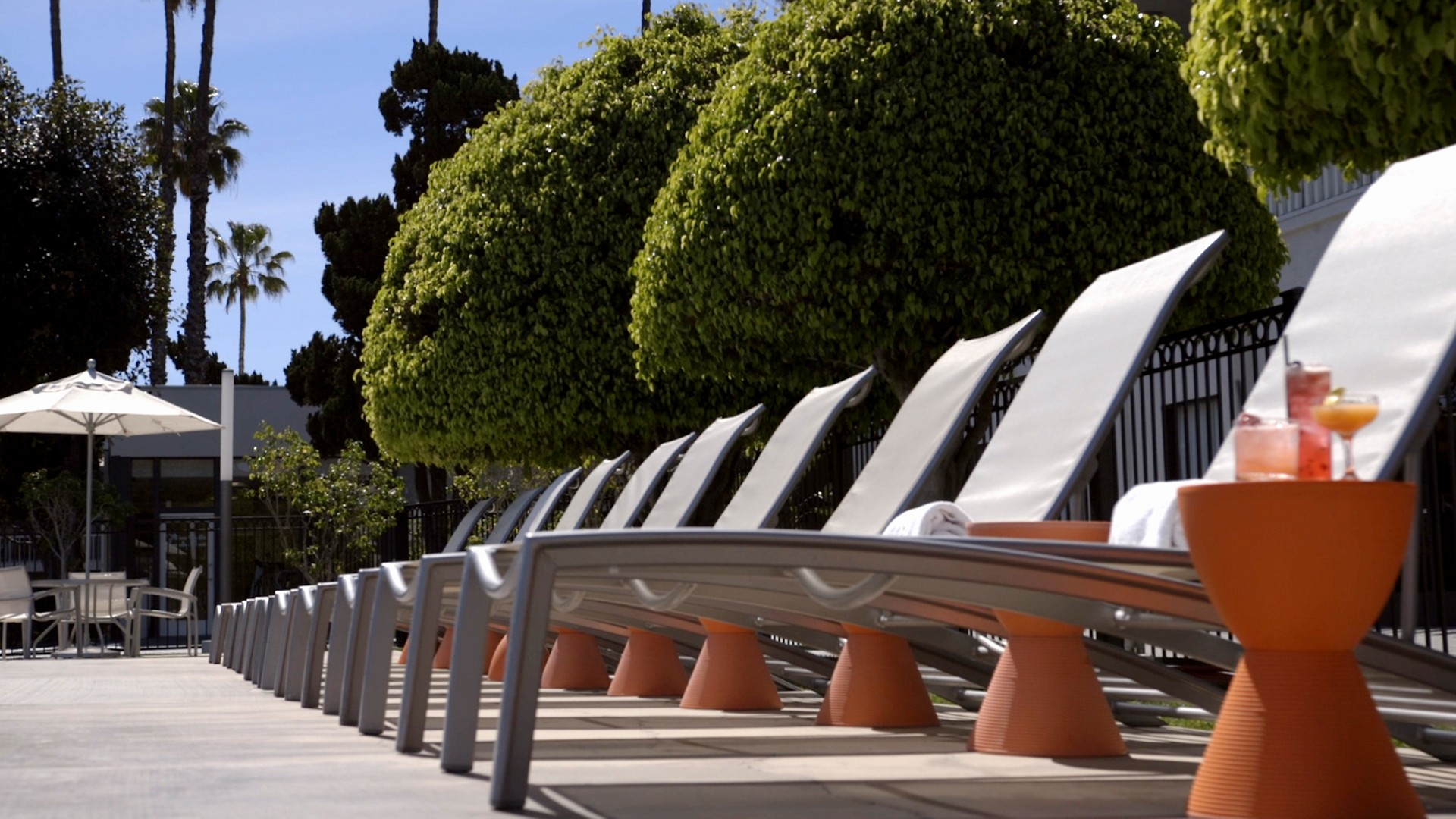 A row of lounge chairs and tables by the pool area with trees and fencing in the background.
